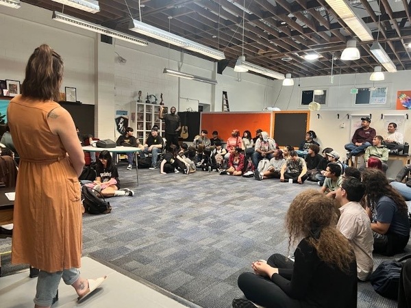 IVA students gathered in horseshoe shape for assembly with teacher standing in front.