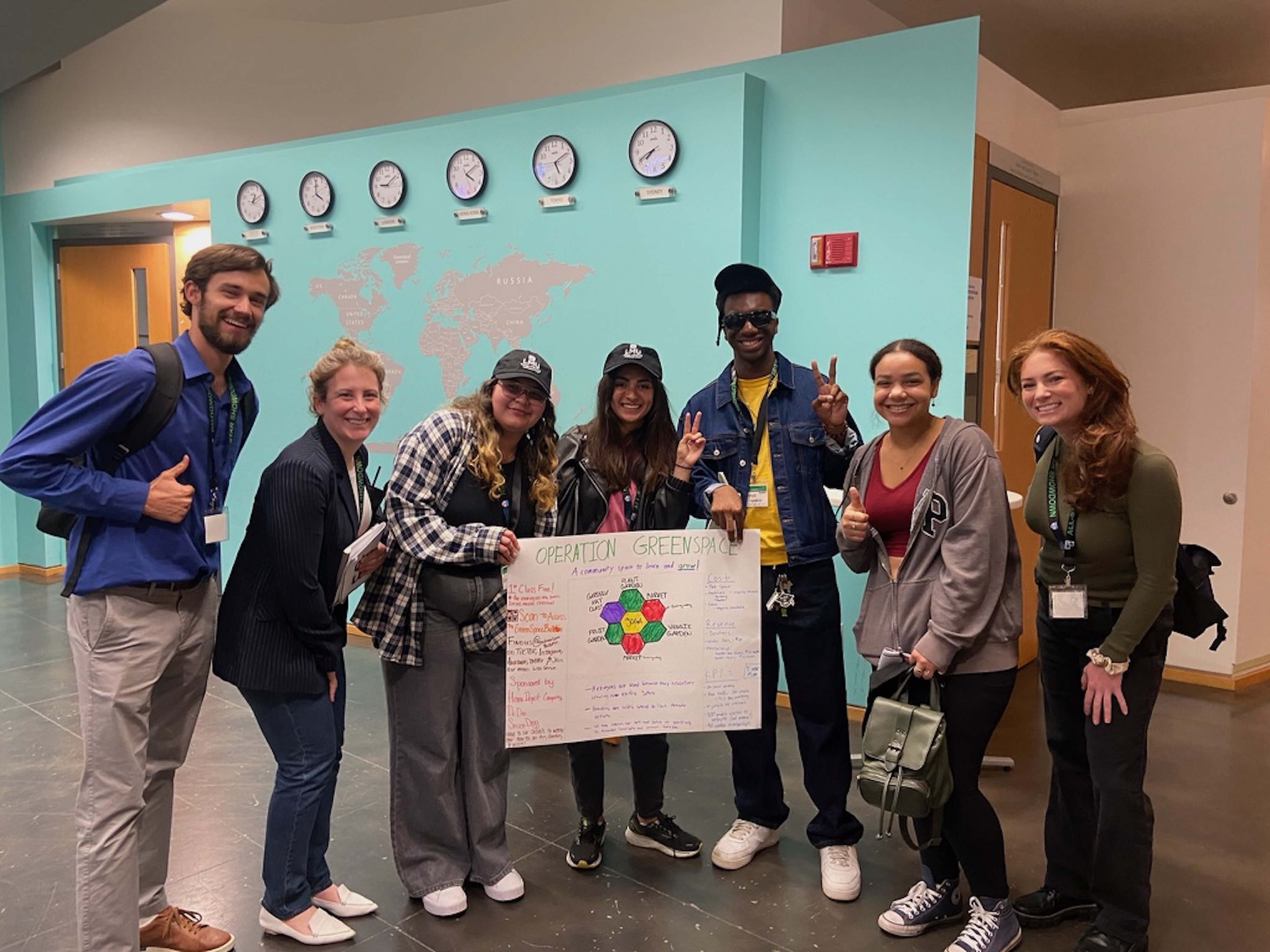 Members of the IVA High community pose together with a poster with the title Operation Greenspace. They are standing in front of a light blue wall with clocks set to world time zones. 